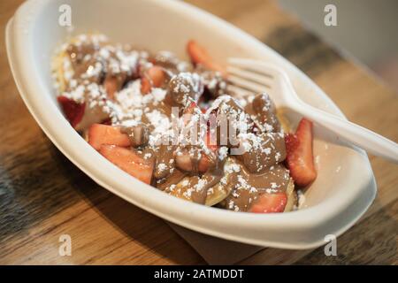 Personne mangeant des poffertjes de crêpes hollandaises avec une fourchette en plastique, poffertjes ont du chocolat, des fraises et du sucre en poudre Banque D'Images