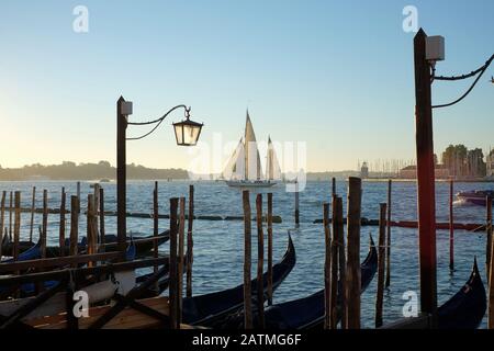 Venise, en Italie, un yacht à deux mâts traverse le bassin de Saint-Marc tandis que des gondoles flottent sur l'eau bleue amarrées à Saint-Marc tôt le matin au bord de l'eau. Banque D'Images