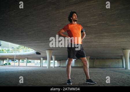 Jeune homme de fitness caucasien se reposant après l'entraînement avec ses mains sur la hanche debout sous le pont - fit un homme prenant un reniflard Banque D'Images