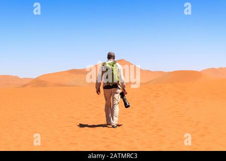 Homme avec sac à dos et grand appareil photo marchant sur les dunes de sable rouge de Sossusvlei, Namib Naukluft Park, Namibie Banque D'Images