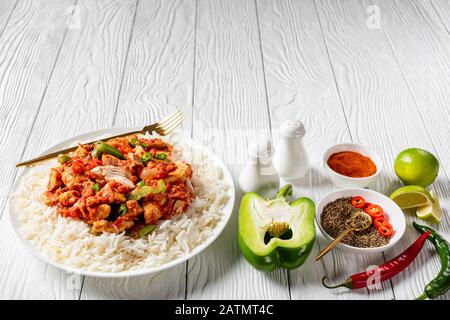 dinde frisées garnies de légumes et sauce tomate surmontée sur riz sur une plaque blanche avec fourchette dorée sur une table en bois avec ingrédients, vie horizontale Banque D'Images