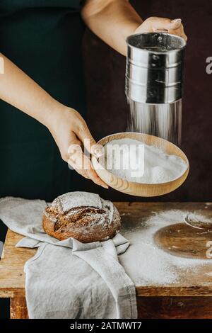 Femme sifant la farine à travers un tamis sur une table en bois. Le chef en tablier sombre sifts la farine à travers un tamis pour préparer la pâte à pain sur un b sombre Banque D'Images