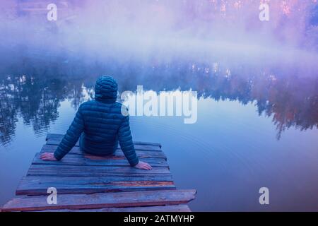 Un homme assis sur une terrasse en bois et regardant un lac avec une rive en granit. Lever du soleil sur le lac Banque D'Images