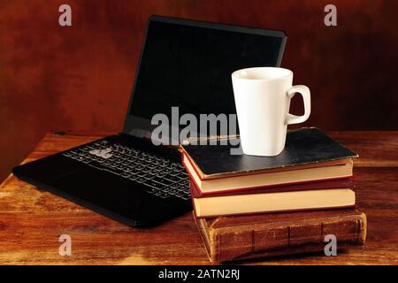 Tasse à café blanche sur la pile de vieux livres anciens. Ordinateur en arrière-plan sur une table marron rayée. Photo horizontale. Banque D'Images