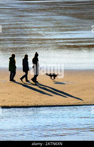 Les gens marchent avec leur chien sur la plage de Porth à marée basse silhouetted par le soleil couchant à Newquay dans Cornwall. Banque D'Images