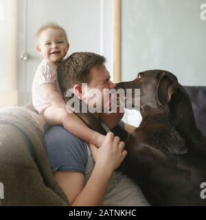 Portrait de famille de jeune beau père avec petit bébé souriant mignon assis sur ses épaules et animal labrador retriever léchant son visage et hugg Banque D'Images