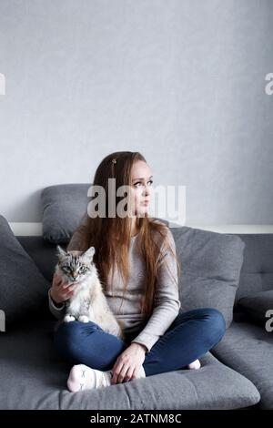 Jeune femme à la mode assise sur le canapé à la maison avec chat domestique. Intérieur gris clair minimaliste. Portrait intégral, espace de copie. Banque D'Images