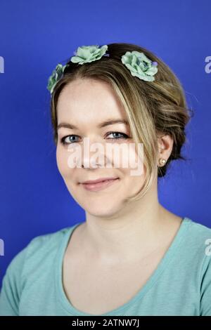 portrait d'une jolie jeune femme blonde caucasienne aux cheveux updo et aux fleurs cyan sur un ciclet de cheveux souriant devant un fond bleu Banque D'Images