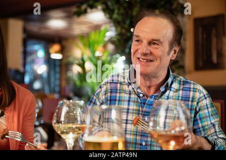 Un groupe d'amis qui dînaient ensemble dans un restaurant, l'accent principal est un homme senior qui mange de la nourriture tout en souriant. Banque D'Images