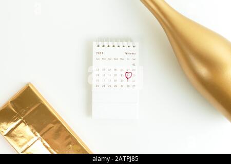 Carte de Saint-Valentin, bouteille de champagne dorée, chocolat dans un wrapper or et calendrier de février avec une date entourée sur une surface blanche. Vue de dessus. Banque D'Images