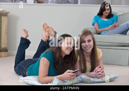 Trois adolescentes à la maison ensemble, deux regardent la télévision et changent de chaîne avec la télécommande et l'autre est dans une chaise d'écoute... Banque D'Images