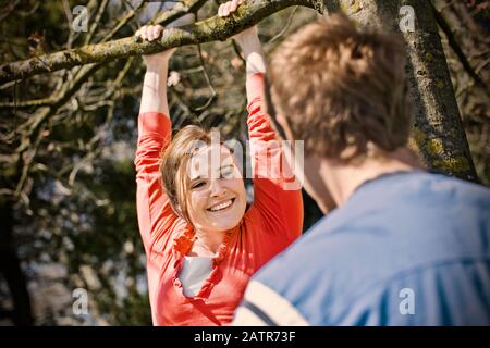 Jeune femme souriante accrochée par ses bras à partir d'un arbre au soleil. Banque D'Images