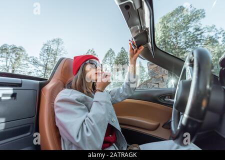 Femme appliquant le maquillage, en tirant sur les lèvres tout en étant assise sur le siège du conducteur de la voiture convertible Banque D'Images