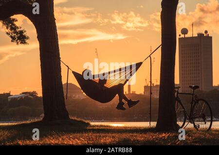 Un jeune homme est assis lisant dans un hamac entre deux arbres au bord de l'eau au coucher du soleil avec un vélo appuyé contre l'arbre et les bâtiments à travers ... Banque D'Images