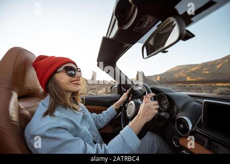 Heureuse femme dans Red Hat conduisant voiture convertible tout en voyageant sur la route du désert avec beau paysage rocheux sur l'arrière-plan pendant un coucher de soleil Banque D'Images