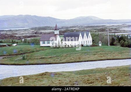Photographie vernaculaire prise sur un film analogique de 35 mm transparent, considérée comme représentant une maison blanche et grise près du lac et du champ vert d'herbe, 1970. () Banque D'Images