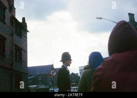 Photographie vernaculaire prise sur une transparence de film analogique de 35 mm, considérée comme un homme dans une veste verte et une casquette noire, probablement un policier, avec des personnes visibles en premier plan, 1970. () Banque D'Images