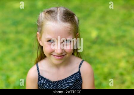 Portrait d'une jeune fille aux cheveux blonds et aux yeux bleus regardant dans l'appareil photo avec l'herbe verte brillante comme arrière-plan; Colombie-Britannique, Canada Banque D'Images