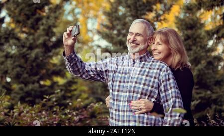 Un couple mature qui profite du temps ensemble et prend un autoportrait tout en admirant le coucher du soleil dans un parc de la ville sur un chaude soirée d'automne Banque D'Images