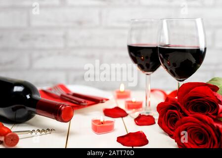 Dîner de Saint Valentin. Deux verres de vin, bouteille de vin, tire-bouchon, roses rouges, assiette et bougies brûlantes sur une table en bois blanc. FO. Sélectif Banque D'Images