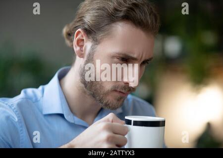 Boire du café tout en méditant. Jeune homme sérieux avec un look élégant avec une tasse de café dans sa main, regardant attentivement avec un sourcil froissé. Banque D'Images