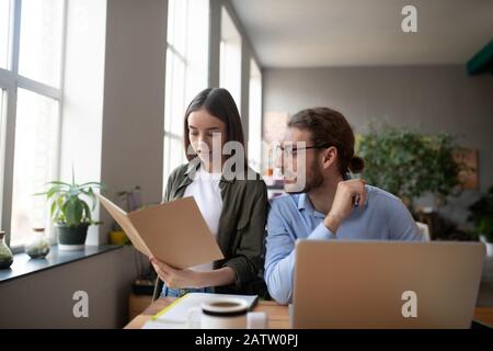 Travailler ensemble. Fille montrant un dossier qui est dans ses mains un homme travaillant sur un ordinateur portable et regardant un dossier, les deux dans une grande humeur joyeuse. Banque D'Images