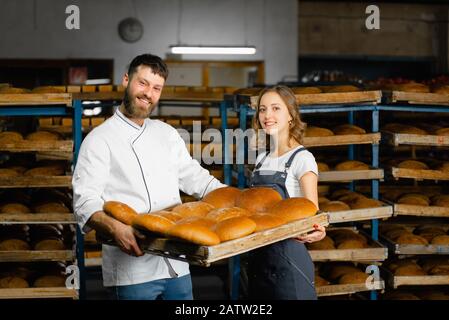 Boulangers à la boulangerie. Un homme et une femme tiennent un plateau avec du pain chaud frais à l'arrière-plan des étagères avec du pain dans une boulangerie. Pain industriel Banque D'Images