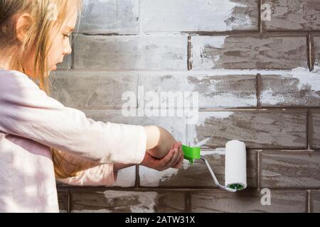 Une petite fille va peindre un mur de briques blanc. Peinture murale, réparations dans la maison. Le peintre, le plâtre peint un mur. Finition des façades avec Banque D'Images