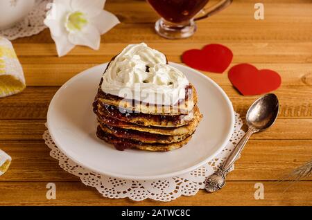 Beignets, crêpes sous forme de coeurs avec confiture et crème fouettée sur un fond en bois clair avec un coeur rouge, un petit déjeuner romantique pour la Saint-Valentin Banque D'Images