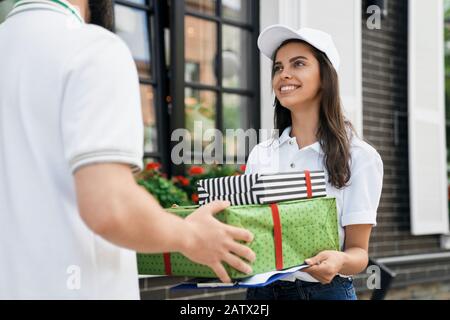 Gros plan de la jeune femme liveuse brunrtte en casquette blanche donnant deux boîtes cadeaux à l'homme. Vue arrière de l'homme incognito dans une tenue décontractée recevant commande et dossier à l'extérieur. Concept de livraison. Banque D'Images