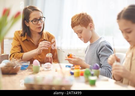 Jeune femme dans des lunettes assis à la table et enseignant aux enfants de peindre les œufs de Pâques Banque D'Images