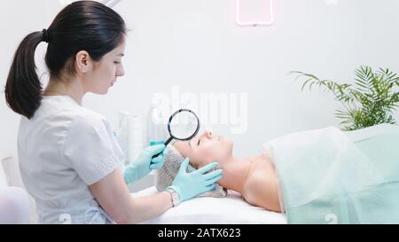 La dermatologue féminine qui examine le visage d'un jeune patient avec une loupe dans une clinique ou un salon de beauté. Concept de nettoyant pour le visage, de peau et de santé Ca Banque D'Images