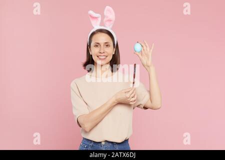 Portrait de belle femme dans les oreilles de lapin tenant l'oeuf et le pinceau et sourire à l'appareil photo isolé sur fond rose Banque D'Images