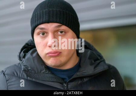 Beau jeune homme avec une expression sceptique soyeuse regardant l'appareil photo avec des lèvres qui pourent dehors en hiver porter un anorak chaud et bonnet beanie Banque D'Images