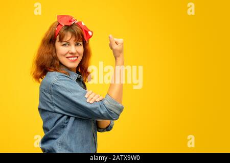 Femme souriante d'âge moyen avec un poing serré qui se déploie sur son manche sur fond jaune, copier l'espace, hommage à l'icône Rosie Riveter Banque D'Images