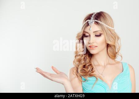 Regardez ici. Femme cheveux blond curly dans tikka bijoux orientaux sur la tête regardant vers le bas souriant montrant avec la main au côté isolé blanc gris arrière-plan Banque D'Images