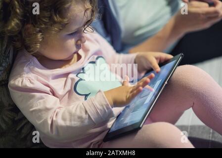 Petite fille se trouve près de sa mère et utilise une tablette numérique. Un enfant de deux ans regarde un appareil électronique à la maison. Banque D'Images