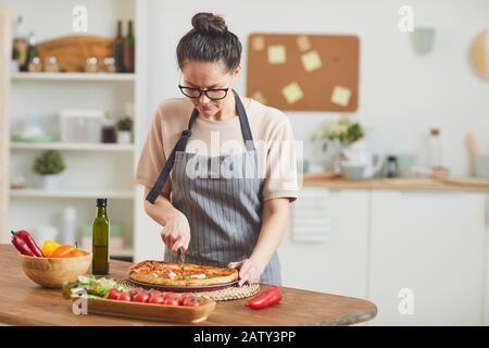 Jeune femme en tablier debout près de la table en bois et couper la pizza maison pour le dîner dans la cuisine à la maison Banque D'Images