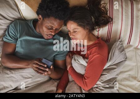 Jeune couple multiethnique allongé sur le lit et en utilisant le téléphone portable ils regardent le film en ligne Banque D'Images