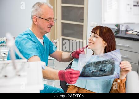 Dentiste souriant de l'homme senior montrant des rayons X dentaires à la jolie patiente, se regardant l'un l'autre, se préparant à un traitement dentaire Banque D'Images