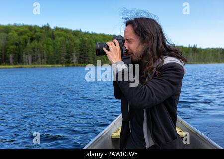 Vue latérale d'un jeune homme d'amérique indigène à poil long qui monte en canoë dans le lac tout en photographiant un paysage pittoresque dans le nord du Québec, au Canada Banque D'Images