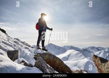 Grimpeur en veste bleue avec bâtons de trekking est debout sur le rocher contre le soleil dans les hautes montagnes enneigées. Conceps d'escalade et d'alpinisme en plein air Banque D'Images