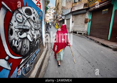 Udaipur, RAJASTHAN, INDE - 03 MARS 2015 : vieille femme dans le foulard rouge qui descend dans la rue avec des graffitis d'art moderne sur le mur dans la ville d'Udaipur. Banque D'Images