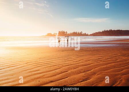 Surfeurs sur la plage marchant vers l'océan. Heure du coucher du soleil, heure d'or. Journée ensoleillée sur la côte de l'océan. Tofino, Colombie-Britannique, Canada Banque D'Images