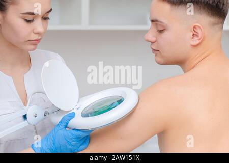 Un dermatologue examine la peau sur le corps d'un jeune homme à travers une loupe d'une lampe de cosmologie. Soins de beauté Banque D'Images