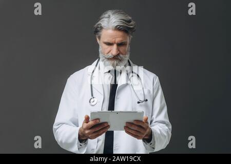 Médecin professionnel de sexe masculin dans un manteau blanc consulter un ordinateur de tablette en lisant l'information avec une expression sérieuse, isolé sur gris bac Banque D'Images