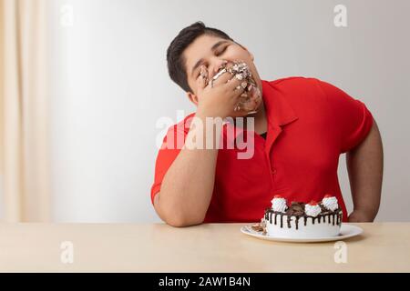 Jeune garçon farce sa bouche avec gâteau. (Obésité) Banque D'Images