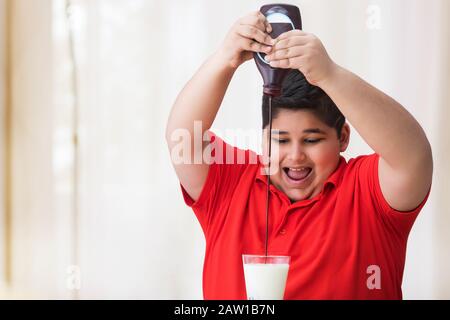 Jeune garçon qui verse du sirop de chocolat dans le verre de lait. (Obésité) Banque D'Images