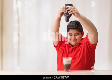 Jeune garçon qui verse du sirop de chocolat dans le verre de lait. (Obésité) Banque D'Images