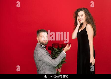 Homme romantique avec bouquet proposant à la petite amie isolée sur fond rouge. Banque D'Images